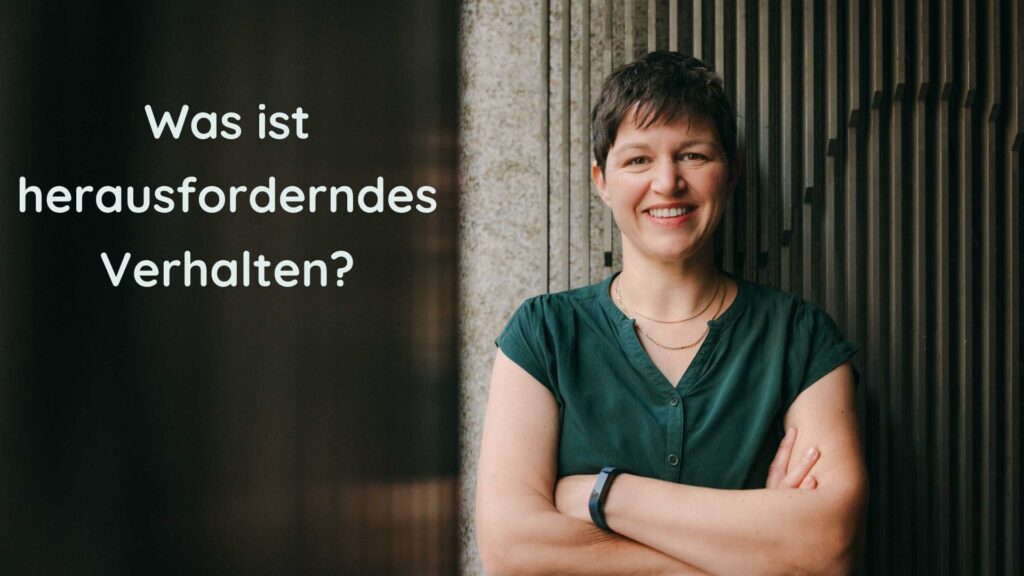 Katja Andergassen, Expertin für herausforderndes Verhalten in Kitas, steht lächelnd mit verschränkten Armen vor einer Wand. Links im Bild steht der Text „Was ist herausforderndes Verhalten?“.