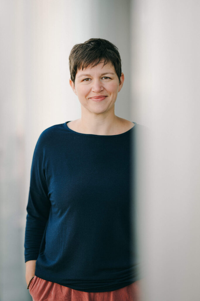 Porträt einer lächelnden Katja Andergassen mit kurzen dunkelbraunen Haaren in dunkelblauem Pullover vor hellem Hintergrund